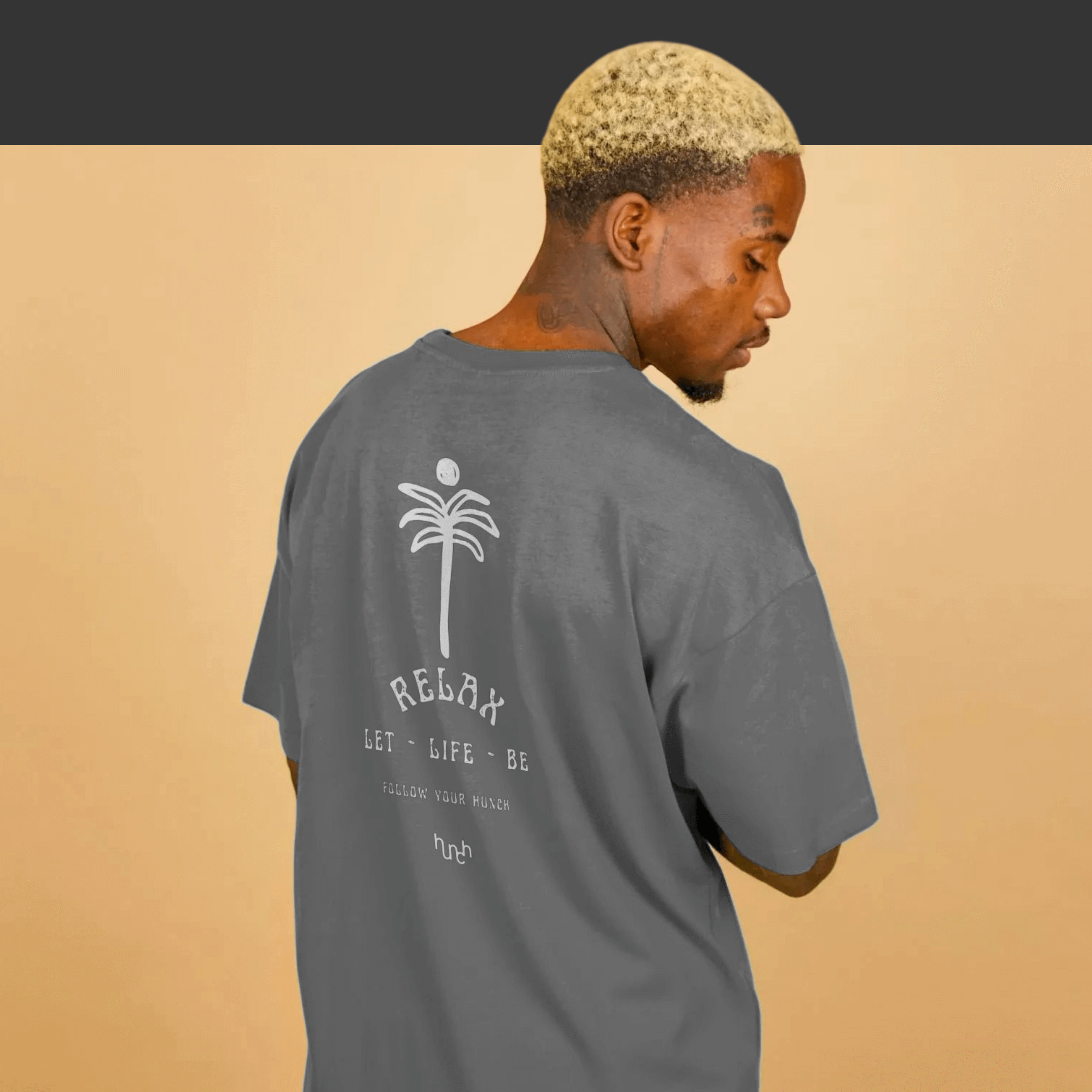 African American man poses with his back turned against a light golden yellow backdrop, showcasing an oversized slate grey Hunch tee with a graphic print ‘Relax’ and a hand-drawn palm tree and sun.