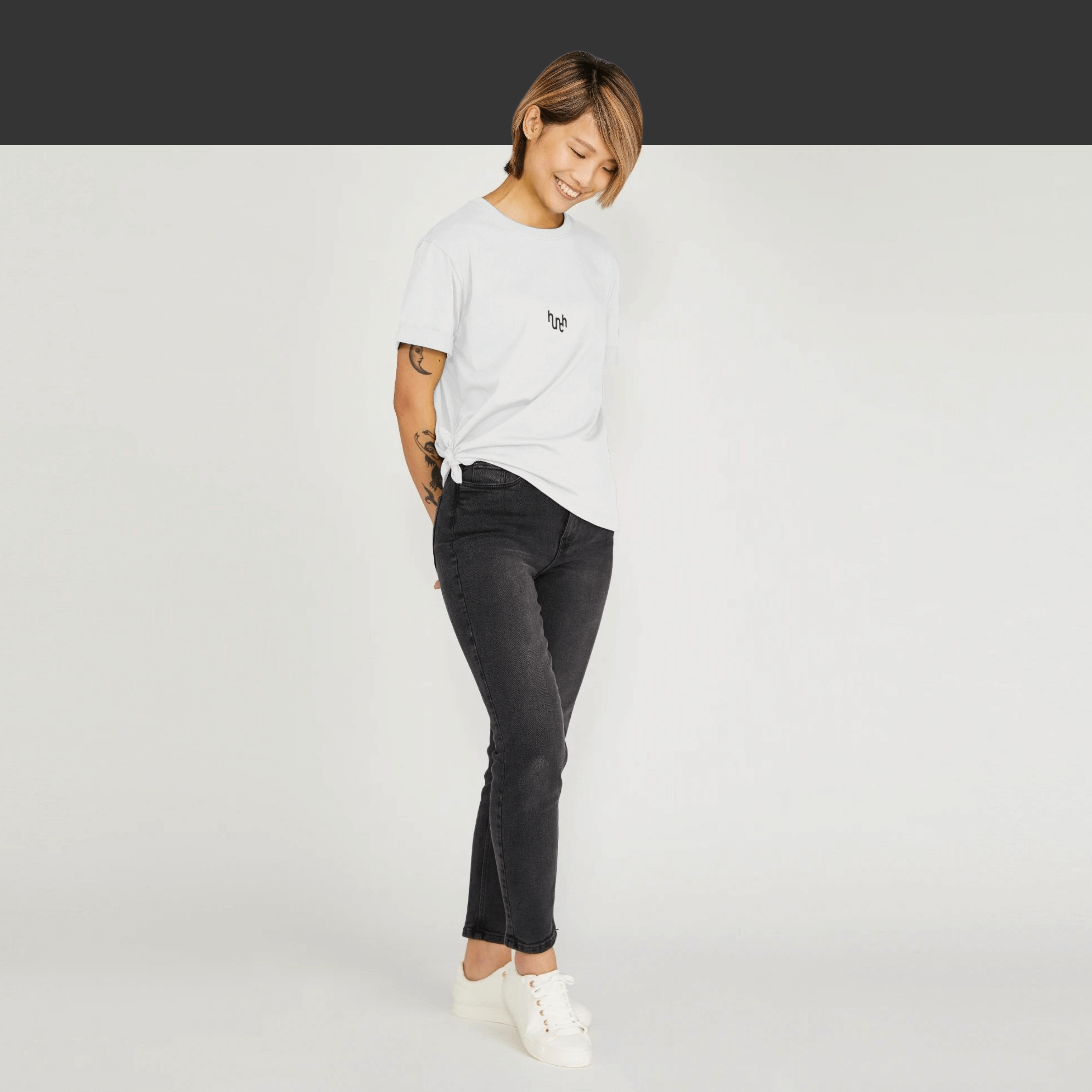 A model of Asian descent stands confidently in black skinny jeans, smiling while wearing a comfortable white recycled cotton tee with a small black centered Hunch logo, against a white background, promoting sustainability.
