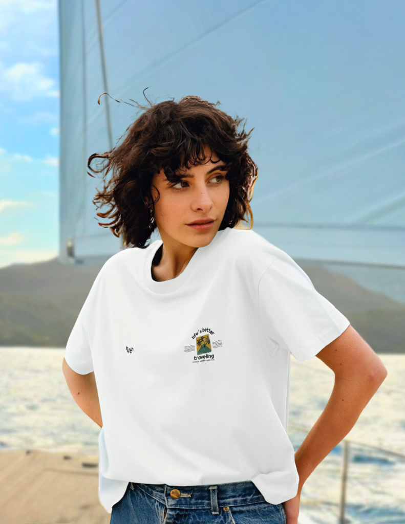 A woman stands sideways on a boat, posing with her hair blowing in the wind, wearing a white oversized 100% organic cotton tee, with the ocean and sails visible in the background, promoting a sense of adventure
