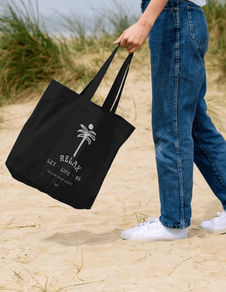 A waist-down view of a woman swinging a black organic tote bag, featuring the ‘Let Life Be’ design, with sea grass flowing behind her as she wears white Hunch barefoot shoes on the beach.